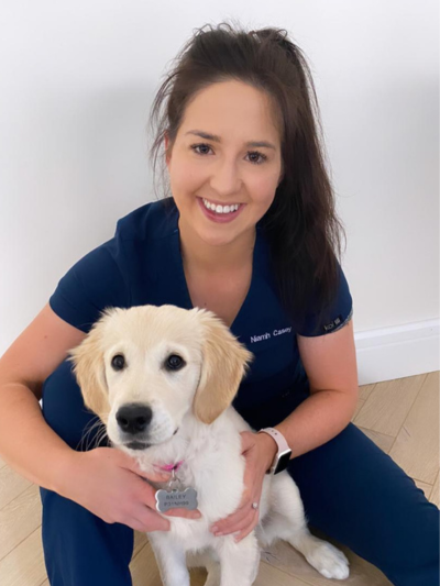 A smiling woman in navy veterinary scrubs, identified by her embroidered name tag as Niamh Casey, poses with a young Golden Retriever puppy named Bailey. They are sitting on a light wooden floor against a plain white wall. The puppy wears a pink collar and a silver name tag.